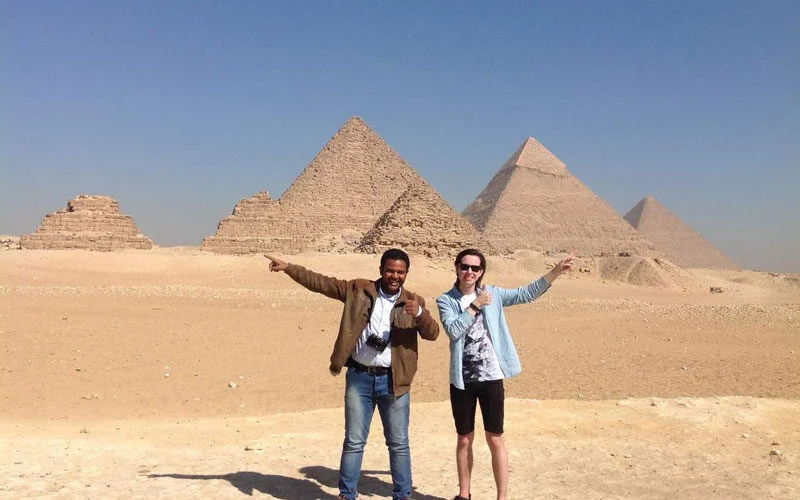 Two men pointing and giving a thumbs up in the desert with several Giza pyramids lined up behind them.