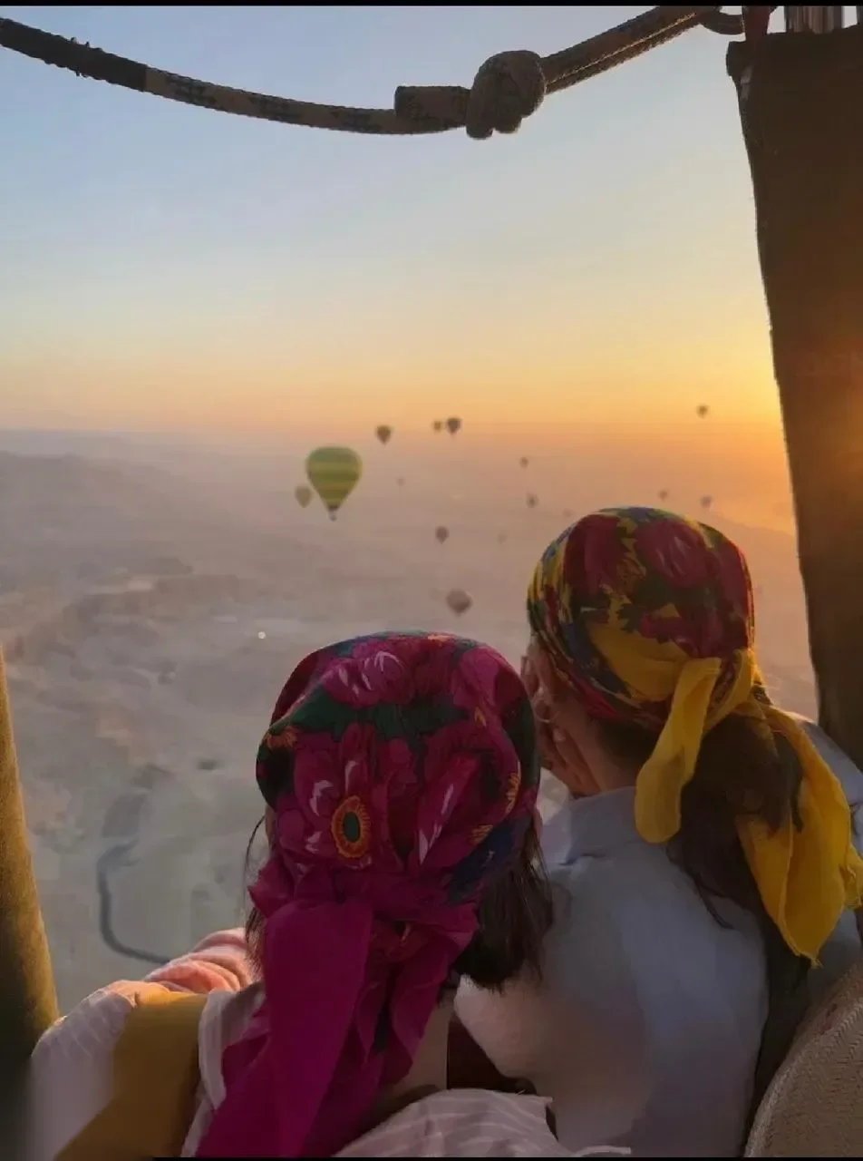 Two passengers in colorful patterned headscarves looking out from a hot air balloon basket at other balloons and the valley