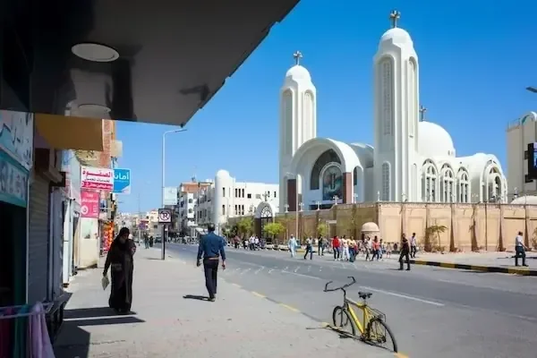 Straße in Hurghada mit den zwei weißen Glockentürmen einer koptischen Kirche mit Kreuzen, daneben Fußgänger und ein Fahrrad