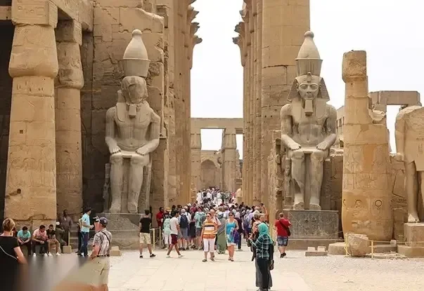 Colossal seated pharaoh statues flank a temple corridor as a large crowd of tourists gathers below