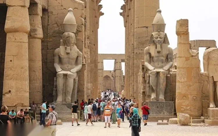 Colossal seated pharaoh statues flank a temple corridor crowded with tourists in white and blue clothing
