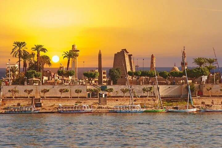 Feluccas are moored along the Luxor riverbank at sunset, with the obelisk and pylon of Luxor Temple visible above the palm