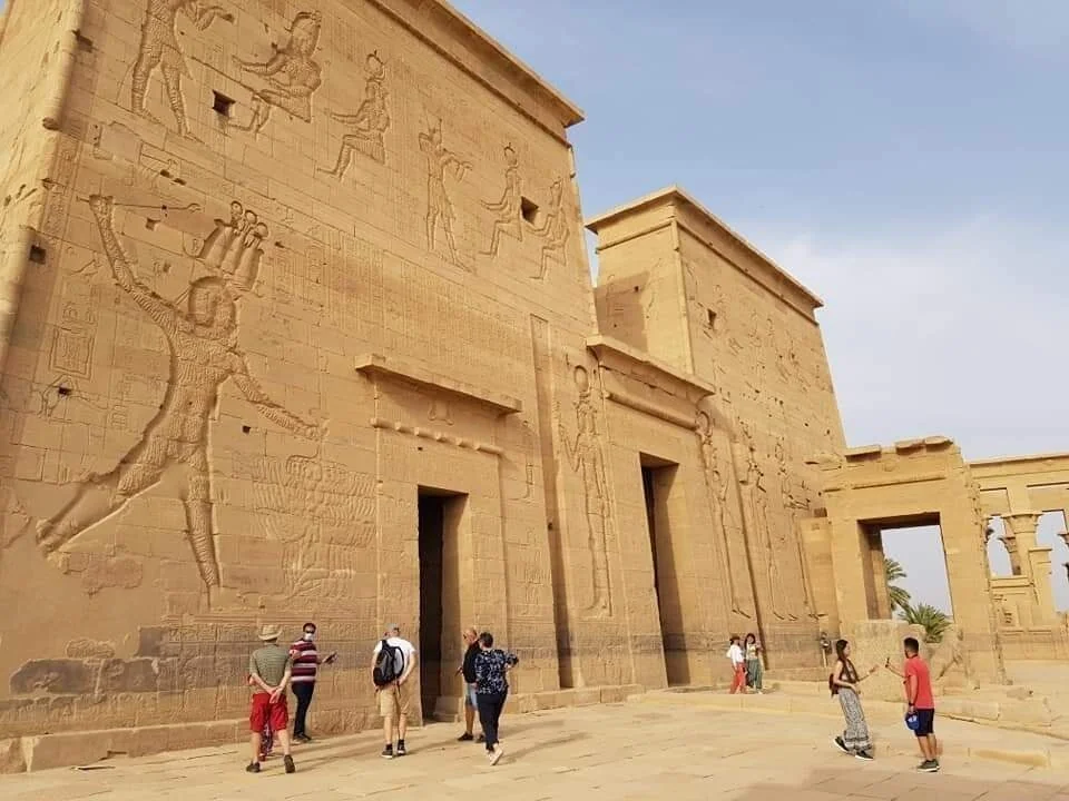 Visitors walk past the carved outer wall of Philae Temple, decorated with reliefs of Egyptian gods and a pharaoh