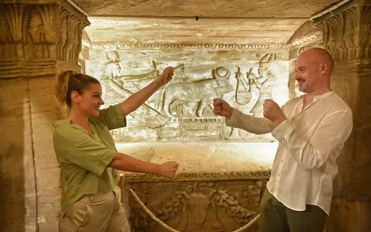 Couple poings levés devant un relief antique de divinités égyptiennes, dans une chambre de catacombe éclairée aux torches