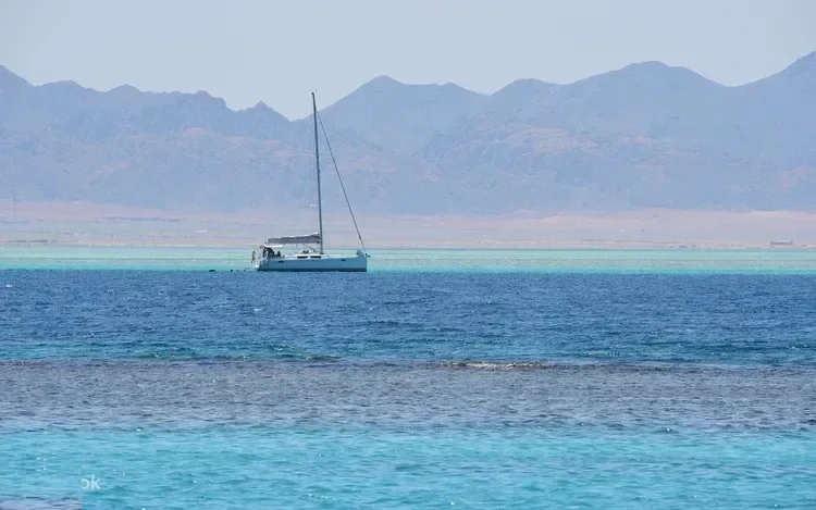 Weißes Segelboot ankert nahe einem Riff, im Hintergrund Wüstenberge an der Küste