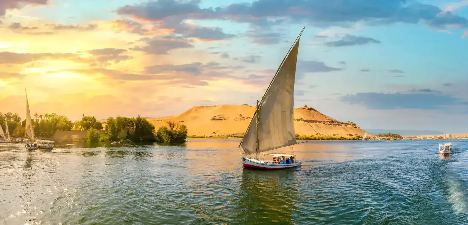 A traditional felucca sailboat with a tall white sail glides on the Nile at golden sunset