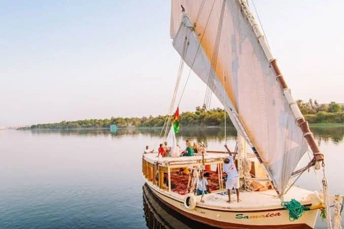 A felucca named 'Samaica' sails calm Nile water with passengers seated on deck and an Egyptian flag flying