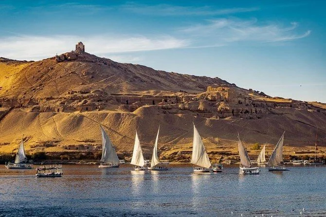 Several white-sailed feluccas glide on the Nile beneath a rocky hill topped with an ancient mausoleum