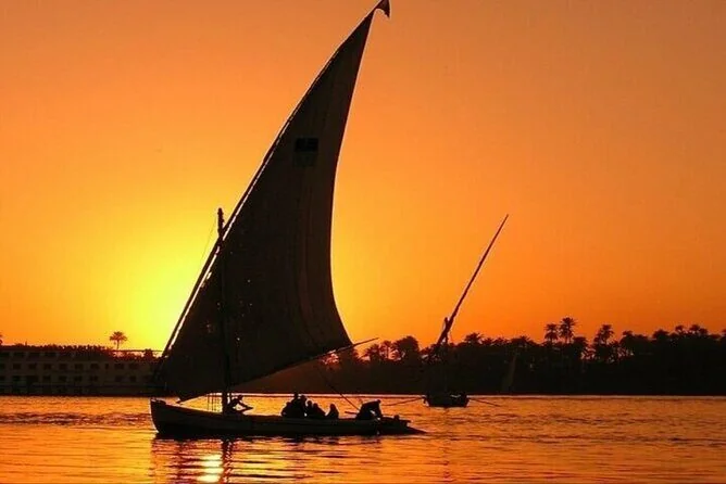 The silhouette of a felucca under full sail against a deep orange sunset sky, palm trees lining the riverbank.