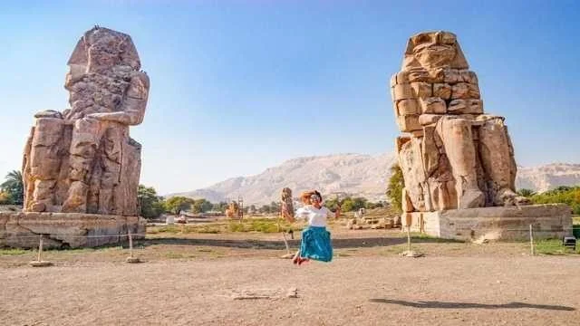 Femme en jupe bleue sautant pour une photo entre les deux colosses de Memnon sous un ciel dégagé