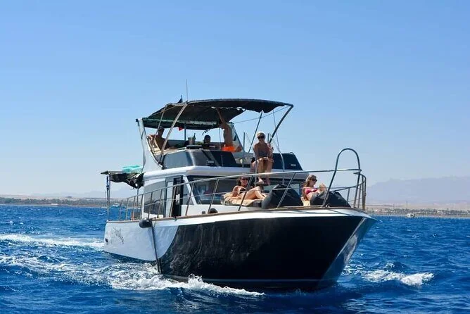 يخت بهيكل أسود يبحر في البحر وركاب يسترخون على سطح التشمّس العلوي والساحل في البعيد
