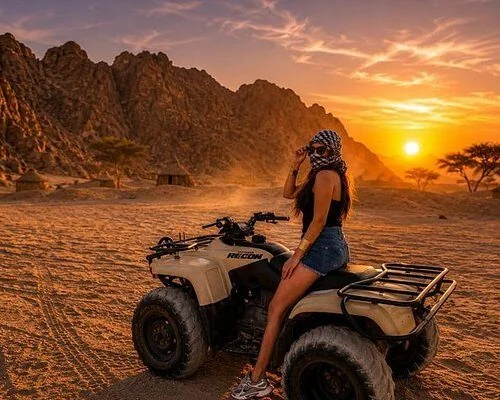 Femme en chèche et short posant sur un quad garé au coucher du soleil, huttes et acacias derrière elle