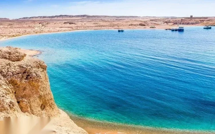 منظر مرتفع لخليج فيروزي منحنٍ بنتوء صخري وشاطئ رملي وقوارب صغيرة راسية في البعيد
