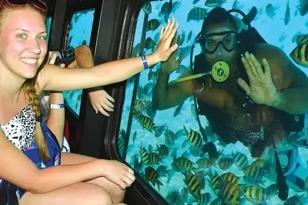 Gast am Unterwasserfenster lächelt und gibt einem Taucher ein High Five, umgeben von einem Schwarm gestreifter Sergeantfische