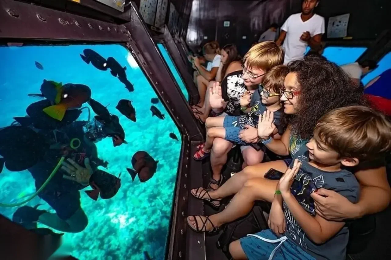 Group of children and a woman waving through an underwater window at a scuba diver surrounded by dark silhouetted fish