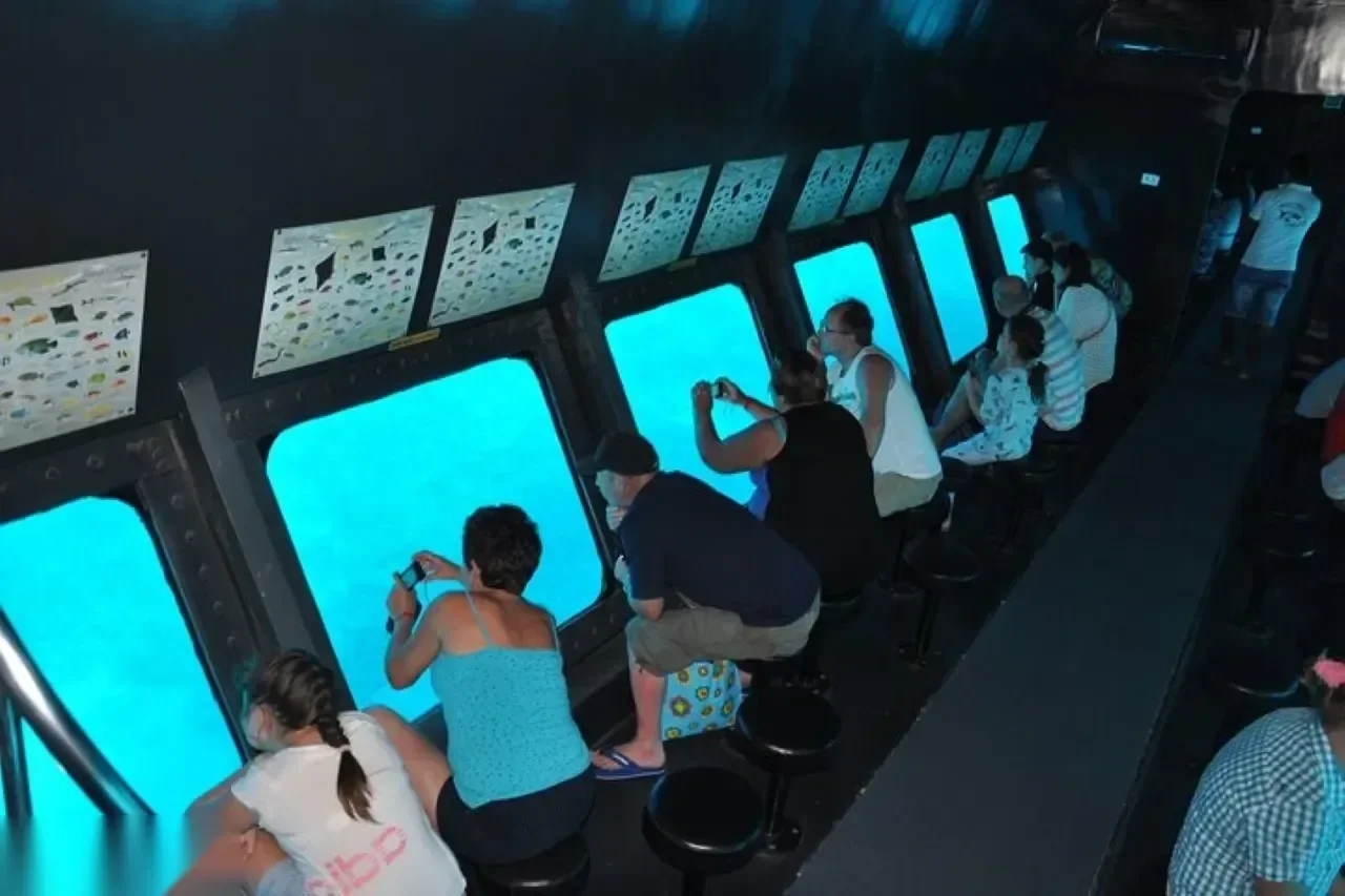 Interior of the semi-submarine's underwater viewing gallery with tourists seated on stools photographing glowing blue windows