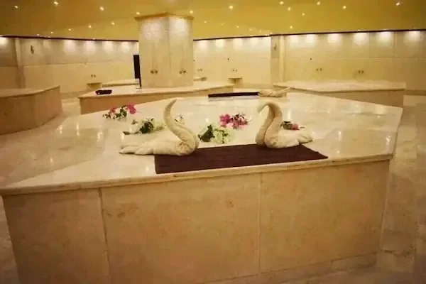 Frau mit Handtuchturban liegt auf einer beheizten Marmorbank neben einem Steinbecken unter einem klassischen Relief im Hammam