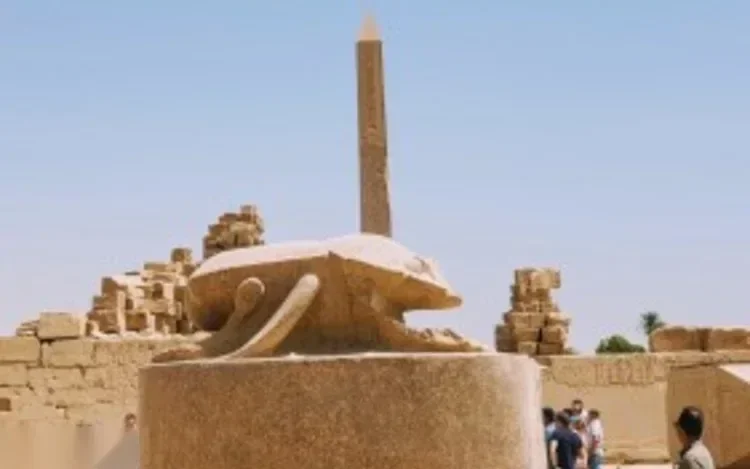 Antike steinerne Skarabäus-Statue auf einem Granitsockel, dahinter hoher Obelisk und Tempelruinen im Karnak-Tempel