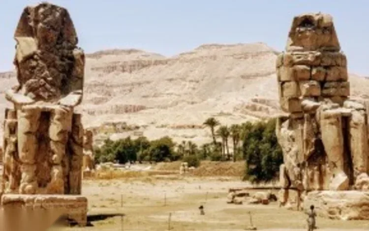Zwei mächtige sitzende Steinstatuen, die Memnon-Kolosse, stehen allein in der Wüstenlandschaft vor Bergen