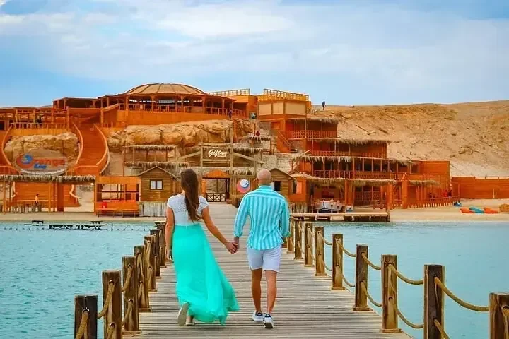 زوجان يمشيان يدًا بيد على رصيف خشبي نحو منتجع شاطئي مبني فوق مياه فيروزية