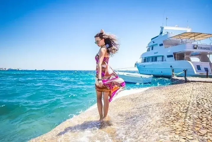 امرأة بفستان ملون منسدل تمشي على رصيف حجري بجانب يخت راسٍ باسم Tiramisu