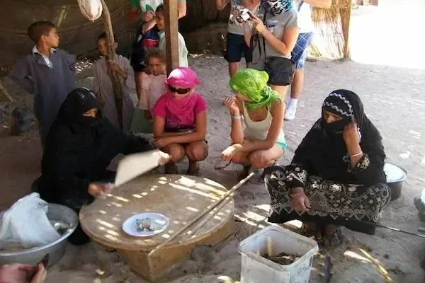 在稻草棚里，贝都因妇女在矮木桌上磨谷物、准备面包，游客在一旁观看