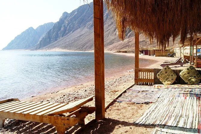 Bains de soleil en bois sous une paillote sur une plage rocheuse au bord d'une baie turquoise calme, montagnes derrière