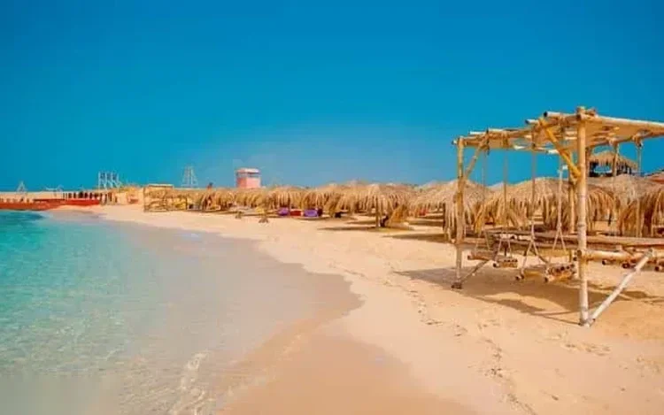 شاطئ رمال بيضاء فارغ بمظلات قشية ومظلة خشبية من الخيزران بجانب مياه فيروزية ضحلة