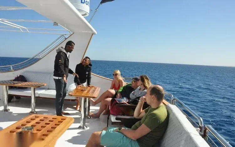 Guests relaxing on a yacht's shaded upper deck seating area with a backgammon table, calm blue sea in the background