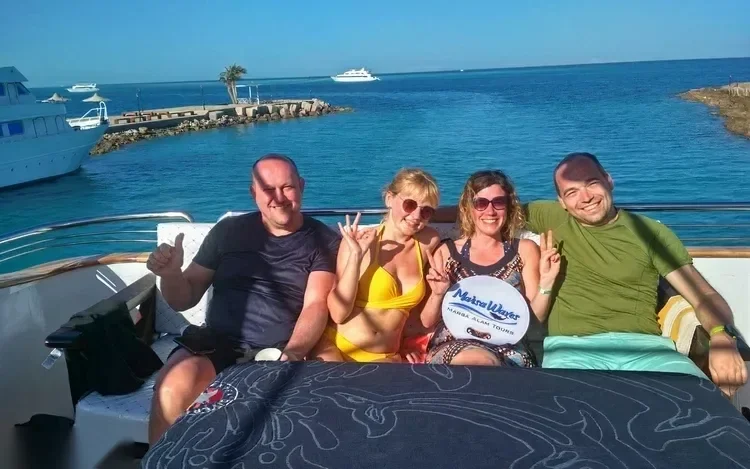 Four friends sitting on a yacht's bow smiling for a photo, harbor with anchored boats and a rock breakwater in the