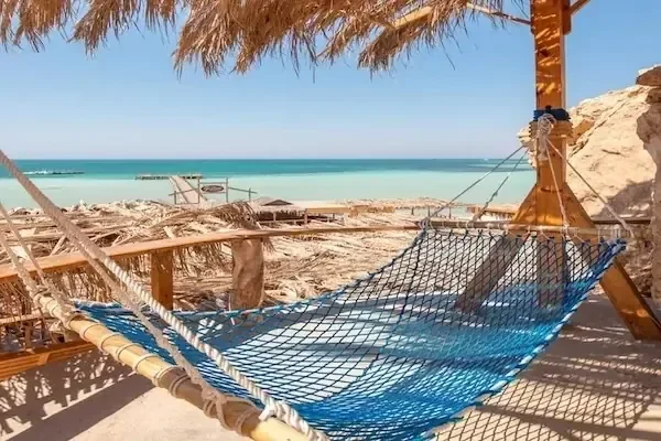 أرجوحة شبكية زرقاء بشبكة حبال معلقة تحت مظلة من القش تطل على خليج فيروزي ورصيف خشبي وأكواخ شاطئ بعيدة