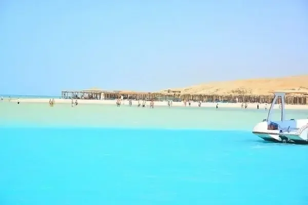 بحيرة فيروزية واسعة وأشخاص يخوضون الماء قرب ساحل رملي تصطف عليه أكواخ القش، وقارب سريع يقترب في المقدمة