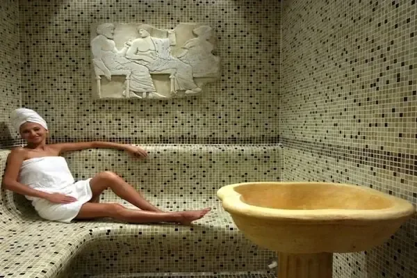 Frau in weißem Handtuch liegt auf einer beheizten Marmorbank im Hammam mit Mosaikfliesen, Steinrelief und großem Steinbecken