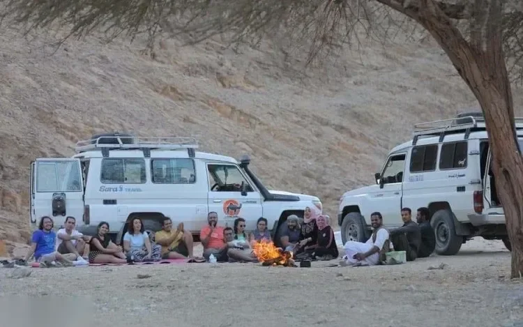Safari group sitting on mats around a campfire beside two white 4x4 jeeps under a large acacia tree