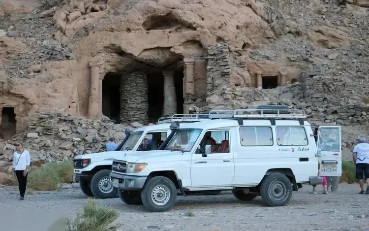 Two white Land Cruiser jeeps parked in front of ancient rock-cut chambers with stone columns carved into a cliffside
