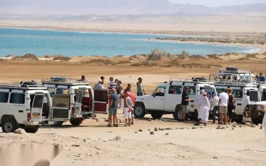 Line of parked safari jeeps and tourists gathered on a bluff overlooking a turquoise coastal lagoon