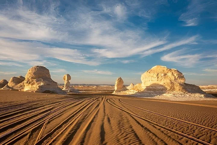Geländewagenspuren im Sand zwischen Kalkformationen unter blauem Wolkenhimmel in der Weißen Wüste