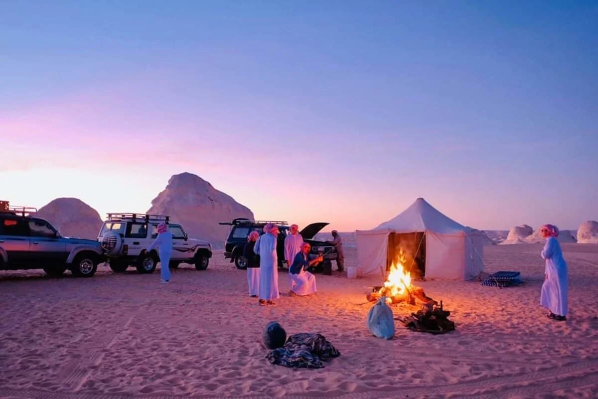 Tentes bédouines, 4x4 et feu de camp sous un ciel rose de crépuscule dans le Désert Blanc