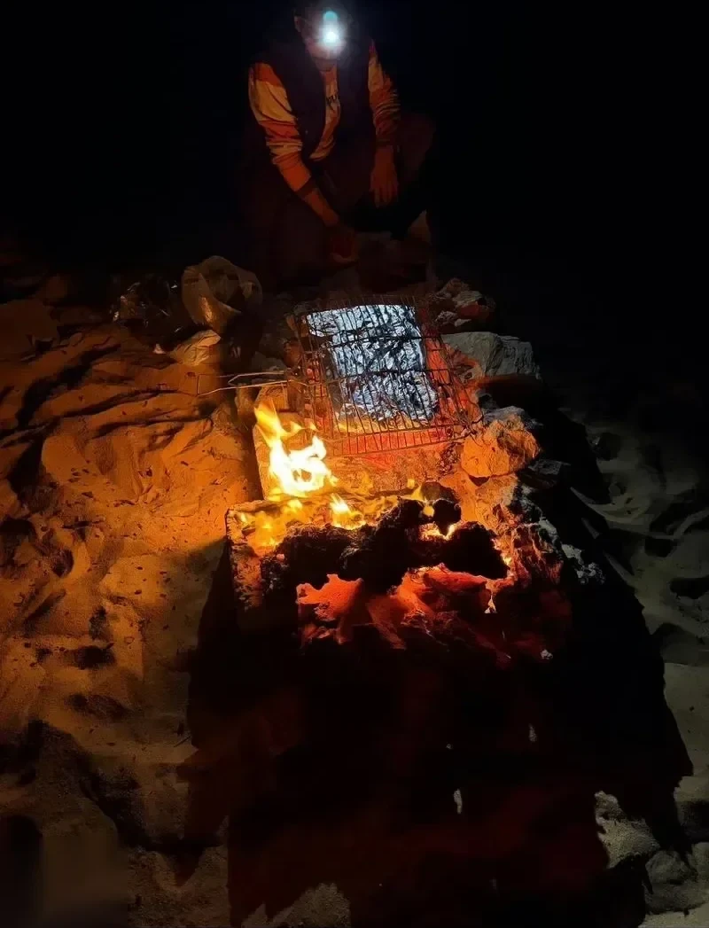Человек в налобном фонаре жарит мясо на проволочной решётке над раскалёнными углями костра ночью в пустыне