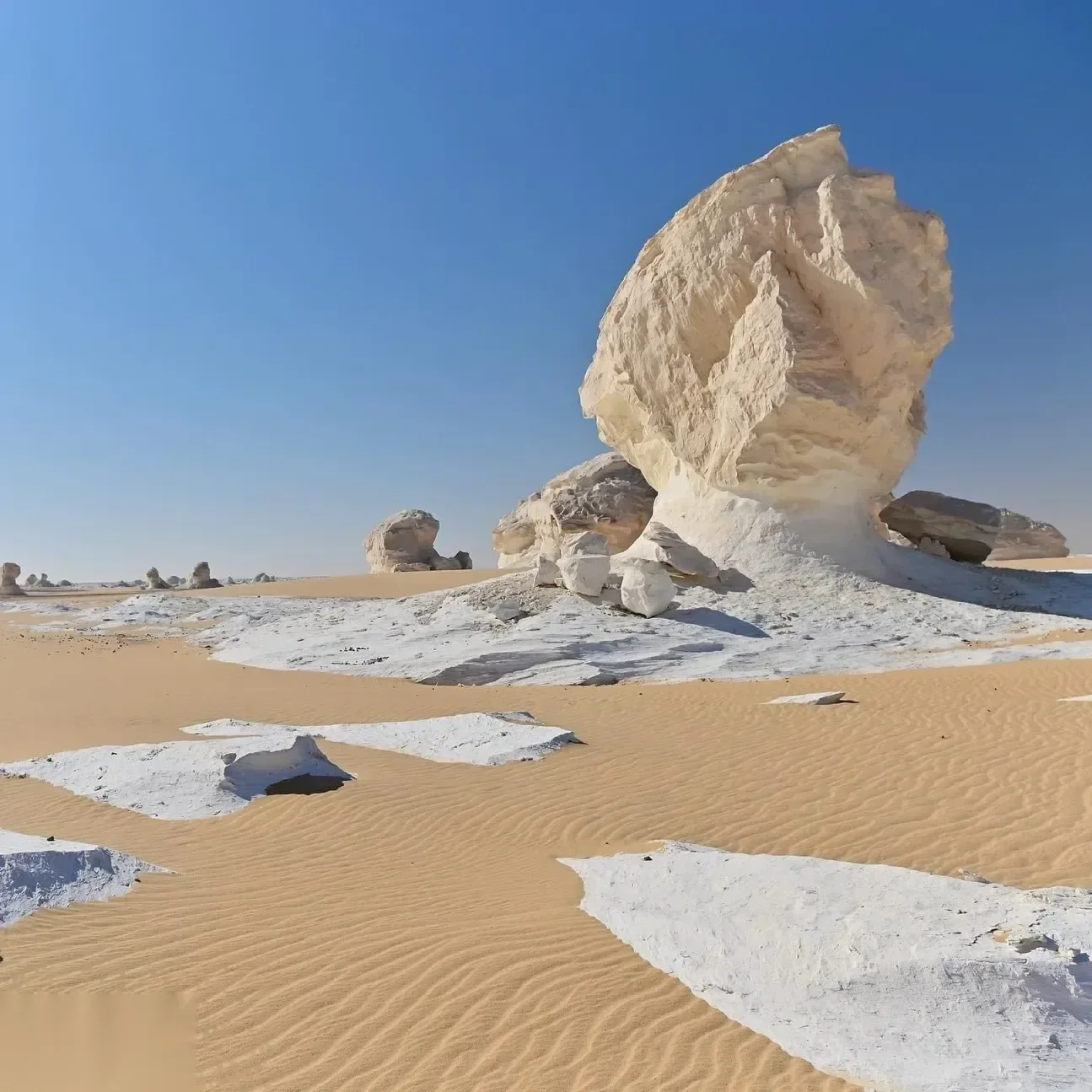 Large white mushroom-shaped chalk rock formation rising from golden sand dunes under a clear blue sky