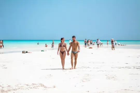 Couple walking hand in hand along a white sand beach beside turquoise water with other beachgoers in the background