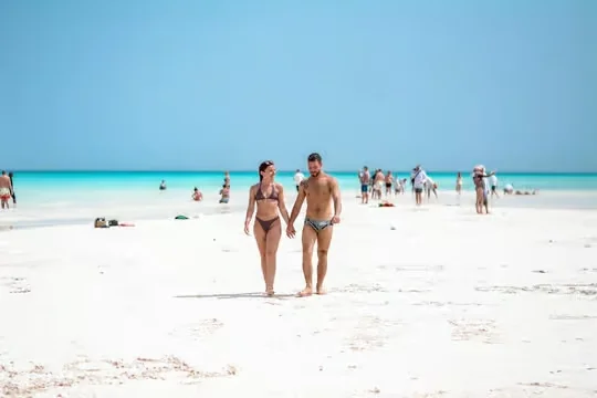 Couple marchant main dans la main sur une plage de sable blanc au bord d'une eau turquoise, d'autres baigneurs au loin