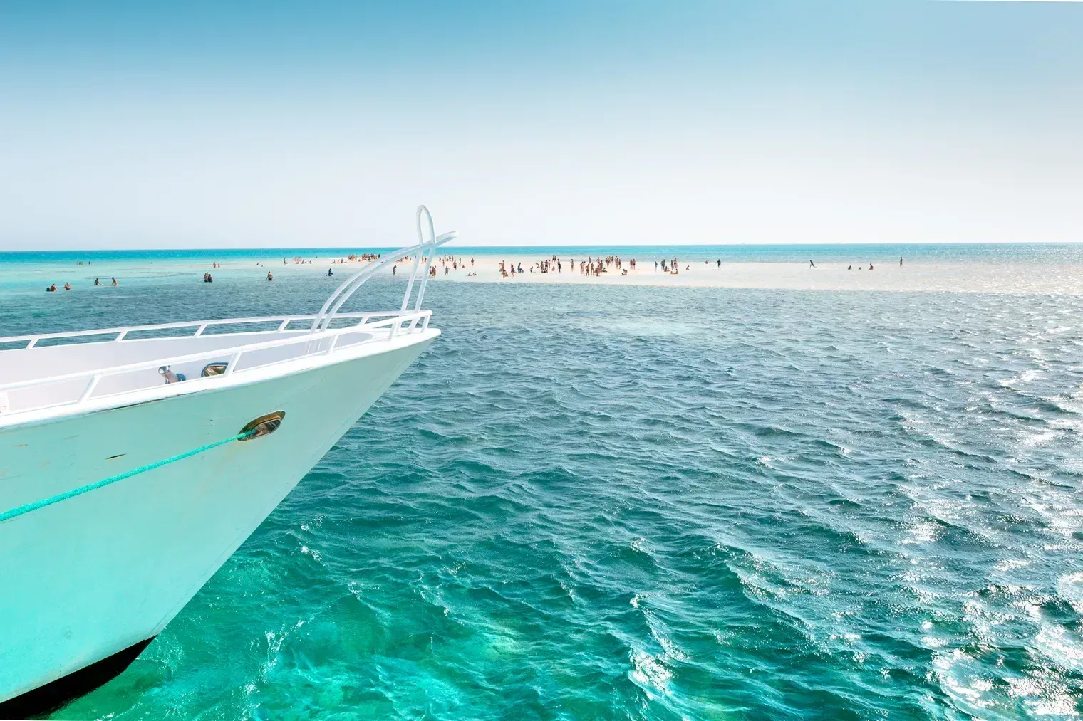Vue depuis l'étrave d'un yacht, une corde turquoise menant vers une île-banc de sable blanc bondée où des baigneurs barbotent