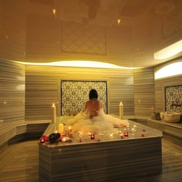 Woman seen from behind sitting wrapped in soap bubbles on a heated hammam platform surrounded by lit candles